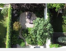 Kleiner, gepflegter Garten aus der Vogelperspektive, mit Kiesfläche, auf der zwei dunkle Liegekissen liegen, umgeben von dichter, grüner Bepflanzung. Rechts befindet sich eine Terrasse mit Holztisch und Stühlen, links und unten von Hecken eingefasst.