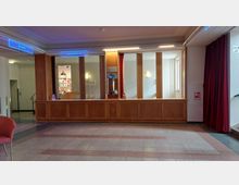 Foyer eines öffentlichen Gebäudes mit einer Holztheke, vertikalen Holzbalken, roten Vorhängen und einem Fliesenboden. Im Hintergrund sind eine Garderobe, leuchtende Wandleuchten und ein Hinweis auf Toiletten sichtbar.