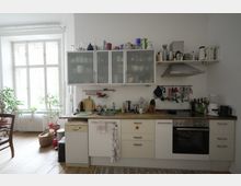 Helle Küche mit einer Arbeitszeile aus Holz, weißen Schränken und Glastüren, darüber offenes Regal und Dunstabzugshaube. Links steht ein großes Fenster mit Pflanzen, Stuhl und Korb auf einem Holzboden.