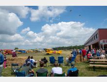 Eine Flugshow auf einem Flugplatz mit Zuschauern auf Stühlen und Heuballen im Vordergrund. Mehrere historische Flugzeuge stehen auf einer Wiese, während zwei Doppeldecker in der Luft fliegen; rechts ist ein Gebäude mit roten Wänden zu sehen.