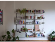 Ein Wohnzimmer mit einer weißen Wand, an der ein hellgraues, offenes Regalsystem befestigt ist. Die Regale sind mit Büchern, Pflanzen, Dekorationsgegenständen und kleinen Figuren gefüllt. Vor dem Regal stehen Zimmerpflanzen auf dem Holzboden. Links befindet sich ein gerahmtes Poster an der Wand.