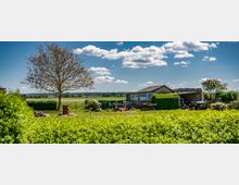 Gartenlandschaft mit einem kleinen, einfachen Gebäude, umgeben von Hecken, Bäumen und einer weiten Wiese im Hintergrund. Im Vordergrund befinden sich ein Feuerplatz, Gartenwerkzeuge und gestapelte Materialien.