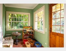 Heller Eingangs- oder Wintergartenraum mit grün gestrichenen Wänden, großen Sprossenfenstern und einer Holztür mit Glaseinsatz. Terrakottafarbener Fliesenboden, kleines Sofa und Tisch sowie ein Regal und Kisten für Gartenutensilien vor Blick ins grüne Außenumfeld.