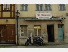 Eine alte, verwitterte Fassade eines historischen Gebäudes mit der Aufschrift „Alte Volksschenke – erbaut um 1743“ über der Eingangstür. Vor dem Gebäude stehen ein schwarzer Motorroller und eine Straßenlaterne, während an den Wänden Graffiti und abblätternde Farbe zu erkennen sind.