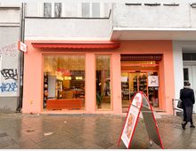 Fassade eines kleinen Geschäftes mit orangefarbener Bemalung und großen Schaufenstern, durch die ein Innenraum mit Regalen und Tischen sichtbar ist. Vor dem Eingang steht ein Werbeschild, und die graue Gehwegplatte davor ist leicht nass.