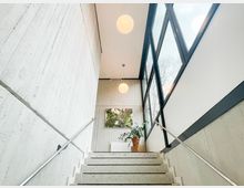 Innenansicht eines modernen Treppenhauses mit Sichtbetonwänden, Terrazzo-Stufen und beidseitigen Metallhandläufen. Rechts sorgt eine hohe Fensterfront mit dunklen Rahmen für Tageslicht; oben hängen runde Kugelleuchten, neben einer Topfpflanze und einem Wandbild am Podest.