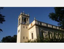 Historisches Gebäude mit einem hohen Turm, verziert mit Säulen und Statuen, im klassizistischen Stil. Im Vordergrund sind Bäume und Büsche zu sehen, während der Himmel klar und blau ist.