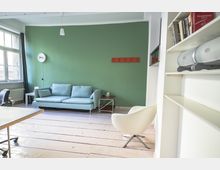 Wohnzimmer mit grüner Wand, einer hellblauen Couch, einem weißen Sessel und einem Bücherregal rechts im Bild. Links steht ein Schreibtisch und ein großes Fenster bietet Tageslicht, während ein heller Holzboden den Raum ergänzt.