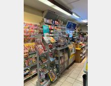 Innenraum eines kleinen Zeitschriften- und Tabakladens mit Regalen voller Magazine, Zeitungen, Zigaretten und Schreibwaren. Im Hintergrund befinden sich ein Lotto-Schalter, Kühlregale mit Getränken und kleinere Verkaufsregale mit Süßwaren.