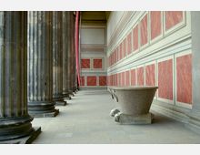 Blick auf eine historische Säulenhalle mit massiven Steinsäulen auf der linken Seite und einer Wand mit rechteckigen, rot-marmorierten Paneelen auf der rechten Seite. Im Vordergrund steht eine große steinerne Wanne auf Löwenfüßen.