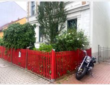 Fassade eines historischen Wohnhauses mit weißen Verzierungen und großen Fenstern, umgeben von einem Vorgarten mit dichter Bepflanzung. Ein roter Lattenzaun und ein geparktes Motorrad auf einem gepflasterten Weg prägen den Vordergrund.