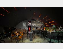 Moderne Bar mit dunkler, gewölbter Decke, beleuchtet durch orangefarbene Lichtleisten. Die Einrichtung umfasst Holz- und Metallstühle, eine lange Theke mit Barhockern, Pflanzen, und Regale mit Flaschen im Hintergrund.