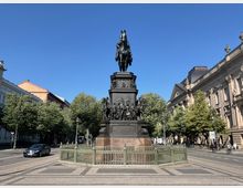 Reiterstatue auf einem hohen Sockel inmitten eines gepflasterten Platzes, von Bäumen und historischen Gebäuden umgeben; im Hintergrund eine klare blaue Himmelsszene. Ein verziertes Metallgeländer umgibt das Denkmal.