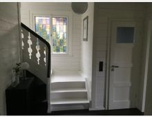 Innenraum eines Hauses mit einer weißen Holztreppe und einem Geländer mit dekorativen Ausschnitten. Rechts befindet sich eine weiße Tür, und im Hintergrund ein Fenster mit buntem, gelbem und blauem Glas. Ein kleiner Schrank mit einer Lampe und einer Vase steht links neben der Treppe.