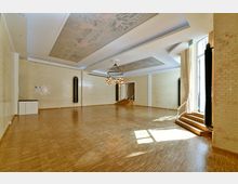 Ein leerer, heller Veranstaltungsraum mit Parkettboden, cremefarbenen Ziegelwänden und großen Fenstern, die von Vorhängen gesäumt sind. Die Decke ist dekoriert und mit einer modernen Hängelampe ausgestattet, während eine kleine Treppe zu einem höher gelegenen Bereich führt.