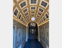 Flur mit reich verziertem, gewölbtem Deckenmuster in Gold- und Beigetönen, kombiniert mit Glasfenstern. Die Wände sind mit dekorativen Wandgemälden versehen, und am Ende des Flurs befindet sich eine aufwendig gestaltete Holztür mit Glas- und Metalldetails.