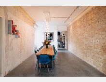 Ein langer Raum mit einer Ziegelwand auf der rechten Seite und einer weißen, leicht abgenutzten Wand links, in der Mitte ein großer Holztisch mit blauen Stühlen und einem Blumenarrangement. Im Hintergrund ist eine minimalistische Küche zu sehen, und von der Decke hängen dekorative Lampen herab.