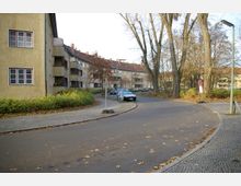 Straßenszene in einem Wohngebiet mit mehrstöckigen, gelblich-beigen Wohnhäusern, pflanzenbewachsenen Vorgärten und vereinzelt parkenden Autos. Der Gehweg ist mit Kopfsteinpflaster bedeckt, und am Straßenrand stehen unbelaubte Bäume.