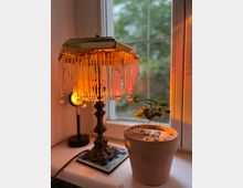 Eine dekorative Tischlampe mit Glasanhängern und warmem Licht steht auf einer Fensterbank neben einem kleinen Terrakottatopf mit grünen Pflanzen. Im Hintergrund ist ein Fenster mit unscharfer Außensicht erkennbar.