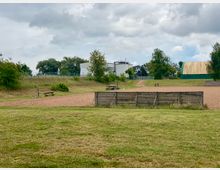 Offene Grünfläche mit einem sandigen Volleyballfeld, umgeben von niedrigen Holzzäunen. Im Hintergrund sind Bäume, ein weißes Gebäude und eine halbrunde grün-gelbe Halle zu sehen, unter einem bewölkten Himmel.