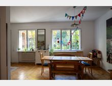 Heller Essbereich mit Holztisch und Stühlen auf Fischgrätparkett, vor großen Sprossenfenstern mit Blick ins Grüne. An der Decke hängen bunte Wimpelgirlanden, daneben stehen eine Kommode mit Spiegel und Zimmerpflanzen.