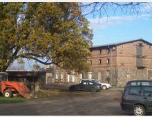 Ein ländlicher Hof mit einem großen Backsteingebäude, das teils aus Feldsteinen besteht, umgeben von geparkten Autos. Im Vordergrund steht ein großer Baum mit herbstlichem Laub, ein Traktor und eine offene Scheune sind ebenfalls sichtbar.