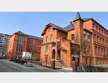 Historisches Backsteingebäude mit mehrstöckiger Fassade, Rundbogenfenstern und einem kleinen Turmaufsatz, daneben ein modernerer Anbau mit großen Fensterflächen. Davor liegen ein gepflasterter Hof mit Zaun, einigen kahlen Bäumen und abgestellten Motorrädern; im Hintergrund ist ein hohes Wohnhaus zu sehen.