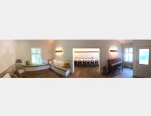 Ein heller Innenraum mit Holzboden, Fenster und Sitzgelegenheiten. Im Vordergrund befinden sich geflieste Sitzbänke mit Kissen und ein Klavier, während im Hintergrund ein Raum mit einer langen Tafel und schwarzen Holzstühlen sichtbar ist.