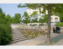 Ein kleiner Park mit einem steinernen Wasserbecken, in dem mehrere große Steine liegen, flankiert von Bäumen und Sträuchern. Drei installationsartige Windradskulpturen aus Metall stehen im Becken; im Hintergrund sind ein Wohngebäude und geparkte Autos zu sehen.