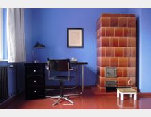 Arbeitszimmer mit blauer Wand, einem minimalistischen Schreibtisch mit schwarzem Stuhl und Tischlampe, sowie einem großen, rotbraun gefliesten Kachelofen auf der rechten Seite. Ein kleiner Holzschemel steht vor dem Ofen, links ist ein Heizkörper unter einem Fenster mit grauem Vorhang sichtbar.