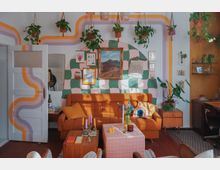 Wohnzimmer mit einer Retro-Einrichtung, geprägt von einer orangefarbenen Couch, bunten Wandmustern in Pastelltönen und mehreren hängenden Pflanzen. Im Vordergrund ein rosa Beistelltisch mit Dekorationen, im Hintergrund Bilderrahmen und Wandteppiche an einer kreativ gestalteten Wand mit grün-weißem Schachbrettmuster.