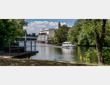 Flussufer mit Bäumen, einem Holzsteg und einer großen weißen Yacht, die auf dem Wasser fährt. Im Hintergrund sind Gebäude am Fluss, eine Brücke und ein Kirchturm zu sehen.