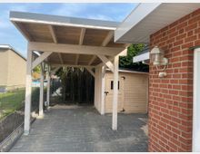 Überdachter Carport aus Holz neben einem roten Ziegelgebäude; Boden aus grauen Fliesen, im Hintergrund ein hölzernes Gartenhaus und ein Zaun.