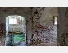 Ein verfallener Raum mit blankgelegenen Ziegelwänden, einem abgenutzten Boden, der teilweise mit grünem Moos bedeckt ist, und einem kleinen Fenster, das schwaches Licht einlässt. Die Wände zeigen deutliche Spuren von Verwitterung, während ein angrenzender Raum durch einen gewölbten Durchgang sichtbar ist.