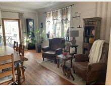 Wohnzimmer mit Holzfußboden, einem Esstisch mit Stühlen links und zwei braunen Ledersesseln rechts. Eine kleine Kommode mit Bücherregal, eine große Standuhr und dekorative Pflanzen schmücken den Raum. Im Hintergrund befindet sich ein Fenster mit Blick auf eine Terrasse. Ein Katze steht zwischen den Sesseln.