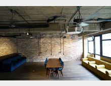 Industrie-inspirierter Innenraum mit einer freiliegenden Ziegelwand, sichtbaren Deckenrohren und metallischen Ventilatoren. In der Mitte steht ein langer Holztisch mit blauen Stühlen, flankiert von einer blauen und einer gelben Polsterbank entlang der Wände.