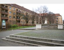 Wohnanlage mit mehrstöckigen Gebäuden im Hintergrund, beige verputzt mit braunen Balkonen, umgeben von kahlen Bäumen. Im Vordergrund ein gepflasterter Platz mit niedrigen Stufen und einem Geländer aus Steinbalustern, daneben ein Grünstreifen und ein Verkehrsschild.