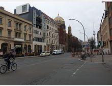 Straßenansicht in Berlin mit verschiedenen Gebäuden. Links befindet sich eine Bar mit Tischen im Außenbereich, in der Mitte die Neue Synagoge mit goldener Kuppel, und rechts im Hintergrund ist der Berliner Fernsehturm zu sehen.