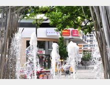 Springbrunnen mit aufsteigendem Wasser in einem städtischen Platz, umgeben von modernen Gebäuden und Geschäften, darunter ein Supermarkt- und Mobilfunkanbieter-Schild; im Hintergrund sind Bäume und eine offene Einkaufszone sichtbar.