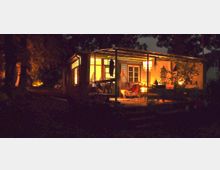 Holzveranda eines kleinen Hauses, bei Nacht beleuchtet mit warmem, gelbem Licht. Die Veranda ist teilweise überdacht, mit Möbeln und Pflanzen dekoriert, während die Umgebung dunkel und von Bäumen umgeben ist.