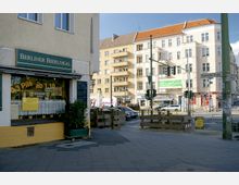 Straßenszene in Berlin mit einer Ecke eines Gebäudes links, das ein Schild "Berliner Bierlokal" trägt, und mehreren mehrstöckigen Wohngebäuden im Hintergrund. Gehwege, eine Baustellenabsperrung und Verkehrsschilder sind sichtbar, dazu Autos und eine Kreuzung mit Ampeln.