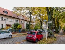 Straßenszene in einer Wohngegend mit Kopfsteinpflaster, zwei Straßenschildern ("Berner Straße" und "Weißdornweg"), geparkten Autos und mehrstöckigen Wohnhäusern mit roten Ziegeldächern. Im Vordergrund stehen Bäume mit Herbstlaub, die den Gehweg und die Straße säumen.