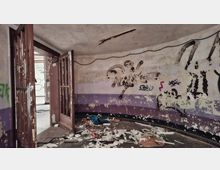 Innenraum eines verlassenen, heruntergekommenen Gebäudes mit abblätternder Farbe, Graffiti an den Wänden und violetten Wandstreifen. Offene, beschädigte Türen führen in einen weiteren Raum; der Boden ist mit Schutt, Papier und Trümmern bedeckt.