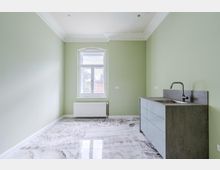 Heller, leerer Küchenraum mit hellgrünen Wänden, hoher Decke mit Stuckleisten und einem großen Fenster über einem Heizkörper. Rechts steht eine moderne Küchenzeile mit grauer Arbeitsplatte, Spülbecken und Wasserhahn; der Boden ist mit glänzenden marmorierten Fliesen ausgelegt.