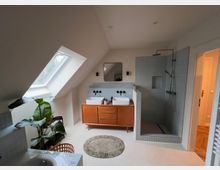 Modernes Badezimmer mit Dachschräge und großem Dachfenster. Der Raum verfügt über ein Doppelwaschbecken auf einem Holzunterschrank, eine offene Dusche mit blauen Wandfliesen, eine grüne Zimmerpflanze und helle Wände.