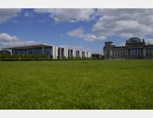 Weitläufige Rasenfläche vor dem modernen Kanzleramt auf der linken Seite und dem historischen Reichstagsgebäude mit Glaskuppel auf der rechten Seite; blauer Himmel mit wenigen Wolken im Hintergrund.