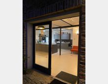 Ansicht durch eine bodentiefe Fensterfront in eine modern ausgestattete Küche mit grauen Schränken, Kücheninsel, Ofen und Deckenbeleuchtung. Der Raum hat helle Fliesen und ist teilweise mit einer roten Sitzgelegenheit möbliert.