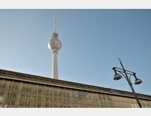 Der Berliner Fernsehturm ragt vor blauem Himmel empor, daneben ein modernes Gebäude mit Glasfassade, in der sich umliegende Strukturen spiegeln, sowie eine schwarze Straßenlaterne im Vordergrund.