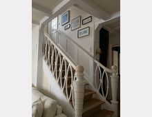 Holztreppe in einem Wohnbereich mit weiß gestrichenem Geländer und dekorativen, ovalen Mustern. An den Wänden hängen mehrere gerahmte Bilder, und im Hintergrund ist eine Tür mit Glaseinsatz sichtbar.