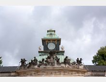 Historische Kuppel eines Gebäudes mit grünlicher Patina, verziert mit schwarzen Skulpturen mehrerer Figuren entlang des Dachrands. Darüber ein Turm mit einem runden Fenster, goldenen Ornamenten und weißen Vasen; bewölkter Himmel im Hintergrund.